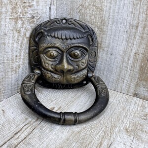 May include: Bronze-colored door knocker featuring a stylized monkey face with a ring handle. The knocker has intricate details and is mounted on a wooden surface. The monkey face has detailed eyes, nose, and mouth.