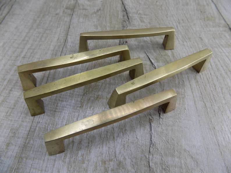 Op de afbeelding: Vijf messing kastgrepen met een rechthoekige vorm en een licht gebogen ontwerp. De grepen zijn in een verspringend patroon op een houten oppervlak gerangschikt.