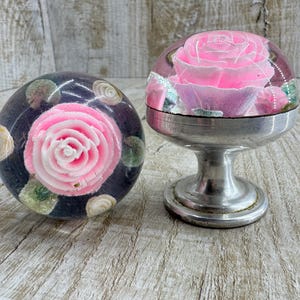 May include: Two decorative glass paperweights. One is a dark blue sphere with a pink rose and seashells. The other is a pink rose encased in a glass dome, mounted on a silver metal pedestal. The items are on a wooden surface.