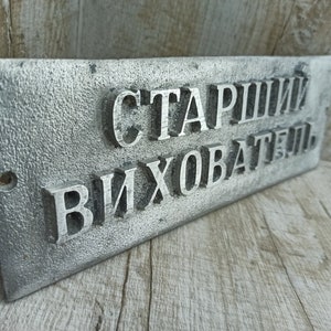 Puede incluir: Un letrero de metal plateado con el texto "Старший Вихователь" en letras en relieve.