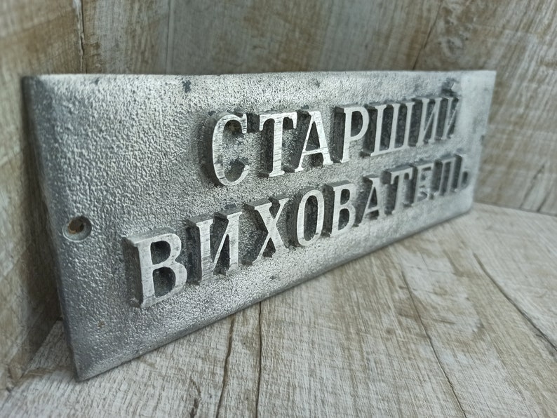 Puede incluir: Un letrero de metal plateado con el texto "Старший Вихователь" en letras en relieve.