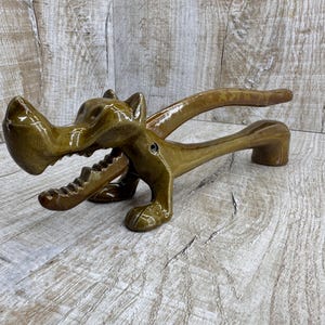 Può includere: Schiaccianoci in ceramica a forma di drago. Il drago, di colore marrone dorato lucido, presenta dettagli come una bocca dentata e zampe artigliate. Lo schiaccianoci ha un meccanismo a leva.