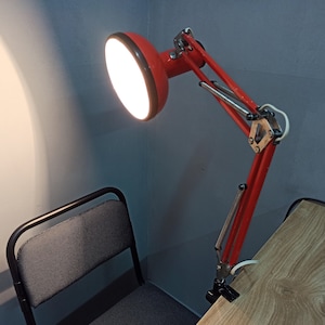 May include: A red adjustable desk lamp with a white light bulb. The lamp is attached to a desk with a clamp.