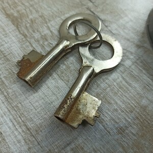 May include: Two antique keys with tarnished metal surfaces and worn brass keyholes. The keys are lying on a light brown wooden surface.