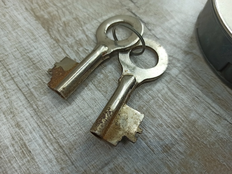 May include: Two antique keys with tarnished metal surfaces and worn brass keyholes. The keys are lying on a light brown wooden surface.
