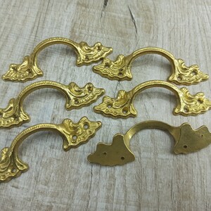 Op de afbeelding: Zeven sierlijke, goudkleurige metalen ladegrepen met een decoratief bloemmotief. Elke greep heeft een gebogen vorm en twee gaten voor montage.