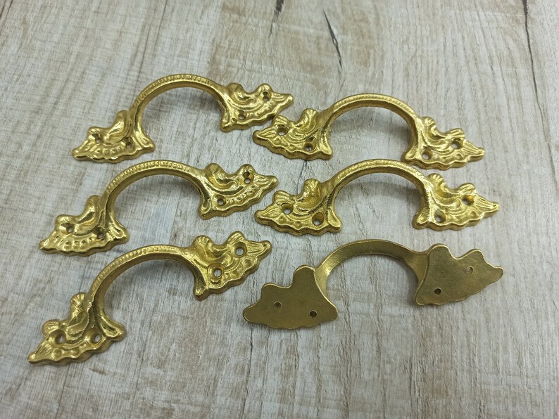 Op de afbeelding: Zeven sierlijke, goudkleurige metalen ladegrepen met een decoratief bloemmotief. Elke greep heeft een gebogen vorm en twee gaten voor montage.