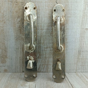 May include: Two antique silver door handles with a rounded rectangular shape and a curved handle. The handles have a worn, aged look with some rust spots.