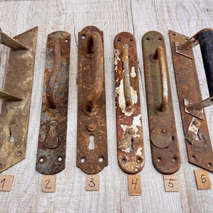 Può includere: Sei maniglie antiche per porte con vari design e livelli di ruggine. Le maniglie in metallo hanno un aspetto rustico e invecchiato. Ogni maniglia è numerata su una piccola etichetta rettangolare.