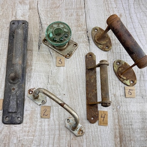 以下が含まれることがあります： 長方形の長い取っ手、緑色のガラス製ドアノブ、さまざまな金属製の引き手とヒンジなど、アンティークのドアハードウェアのコレクション。これらの部品は、古さと使用感を示す、素朴で風化した外観をしています。木製の表面にハードウェアが配置されています。