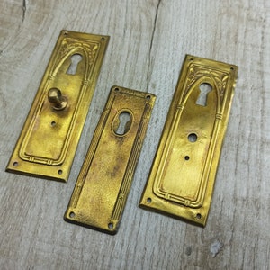 May include: Three antique brass door plates with ornate designs and keyholes. The plates are rectangular and have a slightly aged finish.