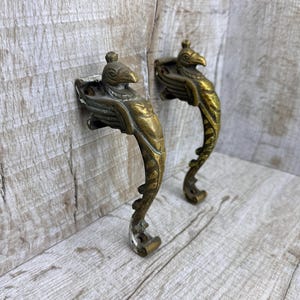 May include: Two antique brass door handles with a bird design. The handles are detailed with wings and a curved, decorative base. The brass has a warm, aged patina, suggesting vintage craftsmanship. The handles are mounted on a light-colored wooden surface.