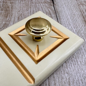 Op de afbeelding: Close-up van een decoratieve kastknop. De knop is goudkleurig en zit op een crèmekleurig oppervlak met gouden geometrische accenten. Het ontwerp heeft een ruitvorm en lineaire elementen.