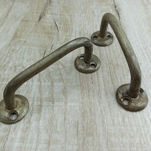 Może przedstawiać: Trzy metalowe uchwyty do szuflad w stylu antycznym z wykończeniem w stylu vintage. Każdy uchwyt ma zaokrąglony, zakrzywiony kształt z dwoma otworami do montażu.