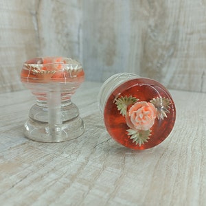 Op de afbeelding: Twee decoratieve kastknoppen met een heldere glazen basis en een rode hars bovenkant. De hars bovenkant heeft een witte bloem met groene bladeren.