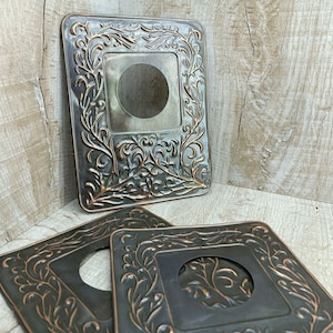 Puede incluir: Tres placas decorativas de metal con forma cuadrada y una abertura circular central. Las placas tienen un diseño floral y de vid ornamentado en color cobre sobre un fondo gris oscuro. Se exhiben sobre una superficie de madera.