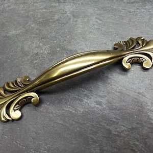 May include: A gold-toned ornate cabinet pull with a decorative scroll design. The pull has a curved shape and is designed for use on furniture.