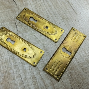 May include: Three antique brass door plates with keyhole cutouts. Two plates have an oval design around the keyhole, while the third has a rectangular shape with vertical lines and a hexagonal keyhole surround. The plates are a warm, aged gold color.