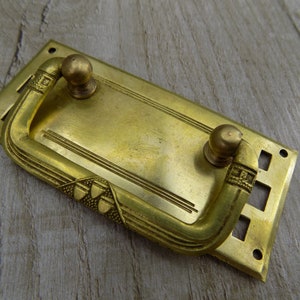 May include: Brass drawer pull with an ornate design. The pull has a rectangular base with a decorative handle that extends out. The handle has two round knobs on either end.