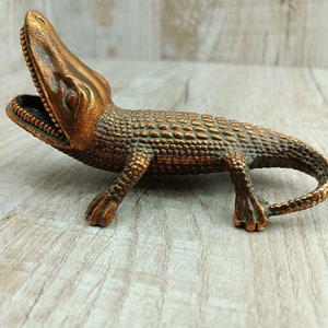 May include: A bronze alligator figurine with an open mouth and detailed scales. The figurine is posed with its head raised and its tail curled.