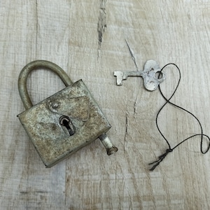 Puede incluir: Un candado de metal oxidado con un ojo de cerradura y una llave plateada atada a una cuerda negra. El candado y la llave están sobre una superficie de madera marrón claro.