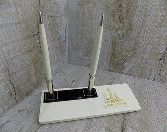 May include: White pen holder with two white pens and a gold design of a mosque on the front. The pen holder is made of plastic and has a black base.