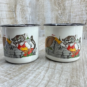 May include: Two white enamel mugs with black rims. Each mug features a cartoon cat in a red vest and yellow pants, holding a sword and standing in front of a castle. The mugs are on a light wood surface.