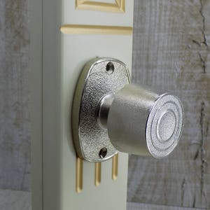 May include: A silver door knob with a round, textured handle. The knob is mounted on a white door with gold accents.