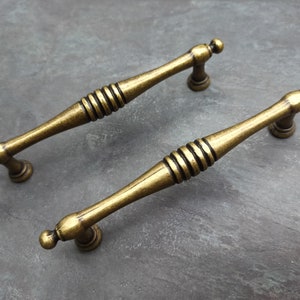 Puede incluir: Dos tiradores de puerta de armario de color dorado antiguo con un diseño acanalado decorativo. Los tiradores son de metal y tienen un estilo tradicional.