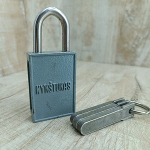 May include: A gray padlock with the word "NYKSTUKAS" embossed on the front. The padlock has a silver shackle and is accompanied by a set of keys.