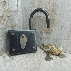May include: A black metal padlock with a silver keyhole and two keys attached to a beige ribbon. The padlock is open and the keys are visible.