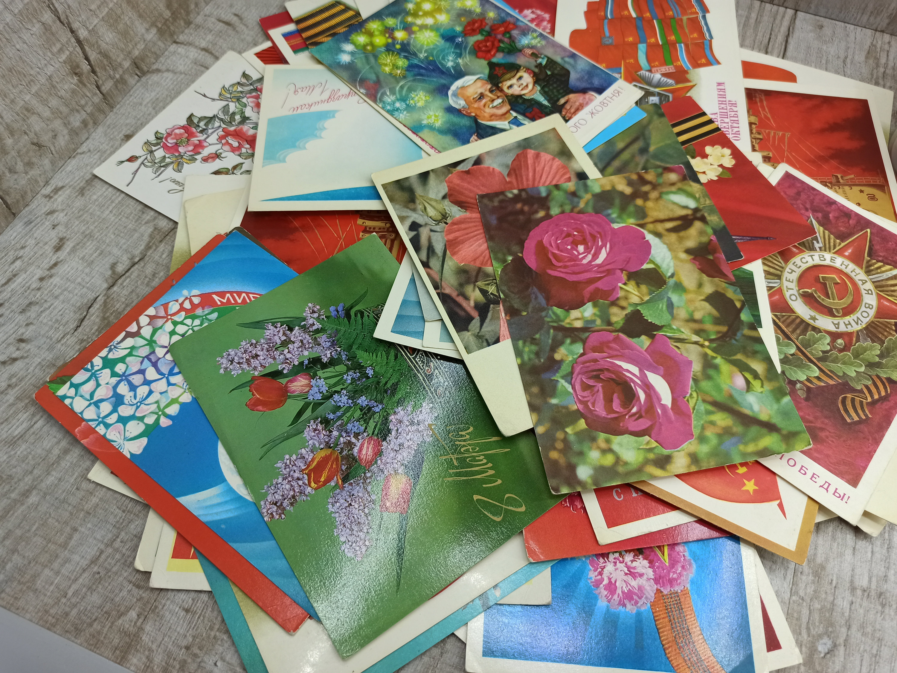 May include: A collection of vintage postcards featuring floral designs, military emblems, and portraits. The postcards display a variety of colors, including red, green, and blue, with some featuring text in a foreign language.