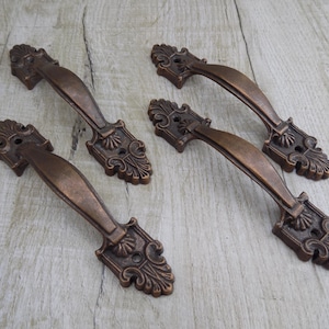 May include: Four ornate, antique-style cabinet door pulls with a copper finish. The pulls have a decorative, scroll-like design and are made of metal.