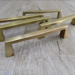 Op de afbeelding: Vier messingkleurige kastgrepen met een modern, geometrisch ontwerp. De grepen zijn in een verspringend patroon op een licht houten oppervlak geplaatst.