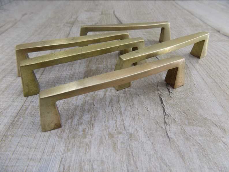 Op de afbeelding: Vier messingkleurige kastgrepen met een modern, geometrisch ontwerp. De grepen zijn in een verspringend patroon op een licht houten oppervlak geplaatst.