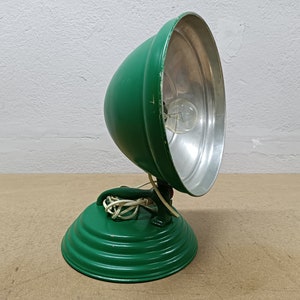 May include: A green metal lamp with a round, reflective shade and a base with a ribbed design. The lamp has a white cord and a light bulb.