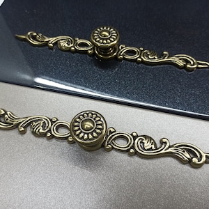 Puede incluir: Dos tiradores de armario de color dorado con un diseño floral decorativo. Los tiradores tienen un pomo redondo con un centro elevado.