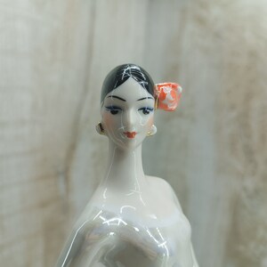 K&ouml;nnte beinhalten: Eine Porzellanfigur einer Frau mit schwarzen Haaren und einer roten Blume im Haar. Sie tr&auml;gt ein wei&szlig;es Kleid und hat einen zarten Gesichtsausdruck.