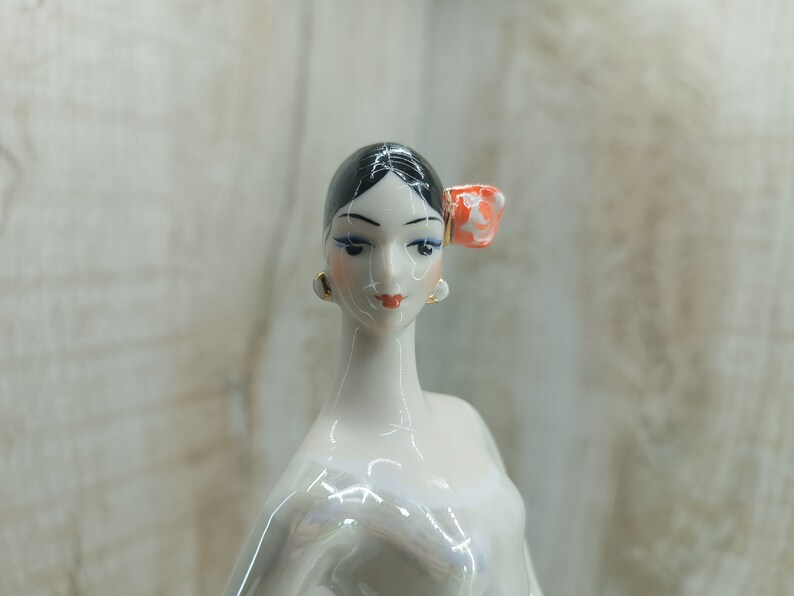 K&ouml;nnte beinhalten: Eine Porzellanfigur einer Frau mit schwarzen Haaren und einer roten Blume im Haar. Sie tr&auml;gt ein wei&szlig;es Kleid und hat einen zarten Gesichtsausdruck.