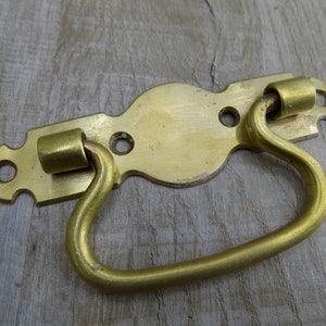 May include: A brass drawer pull with a decorative, ornate design. The pull has a curved handle and two round, decorative accents.