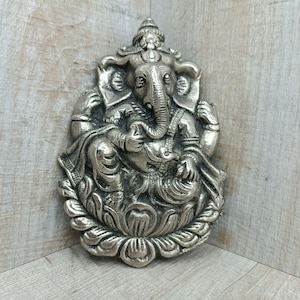 Puede incluir: Colgante de pared de metal plateado de la deidad hindú Ganesha, representado sentado sobre una flor de loto. La escultura está detallada con intrincados tallados y presenta la cabeza y la trompa de elefante características de Ganesha.