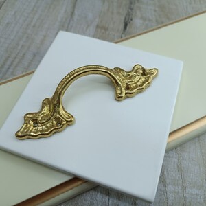 May include: A gold-toned ornate drawer pull with a decorative, leaf-like design. The pull is on a white surface.