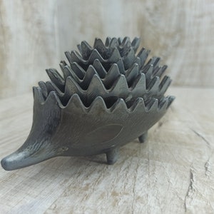 Pode incluir: Objeto em forma de ouriço-cacheiro em metal fundido com acabamento cinza. O ouriço tem um focinho pontudo, pernas pequenas e uma série de espinhos triangulares e serrilhados nas costas. O objeto é provavelmente um item decorativo ou um pequeno recipiente.