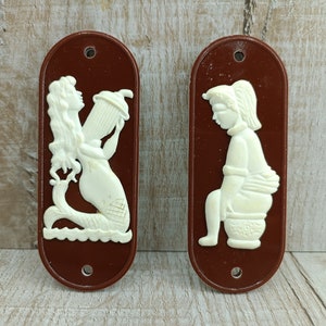 May include: Two vintage bathroom signs with a brown background and white raised figures. The sign on the left features a mermaid holding a shower head. The sign on the right features a person sitting on a toilet.