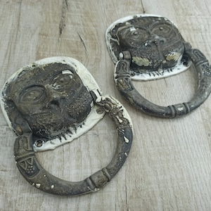 May include: Two antique door knockers with a face design. The knockers are made of metal and have a white painted finish. They are both in the shape of a ring with a face in the centre.