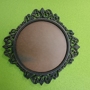 Può includere: Specchio decorativo ornato, color bronzo scuro, con un centro ovale e una cornice elaborata. Lo specchio è posto su uno sfondo verde lime strutturato, creando un contrasto sorprendente. La cornice presenta disegni simmetrici dettagliati.