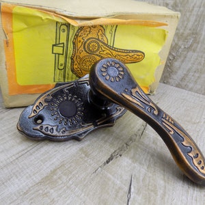 May include: A black and gold door handle with an ornate design. The handle is attached to a plate with a decorative edge. The handle is in a box with a yellow and orange label.