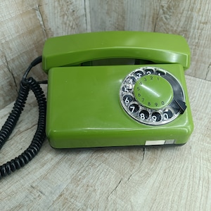 Könnte beinhalten: Ein grünes Wählscheibentelefon mit einem schwarzen Spiralkabel. Das Telefon hat eine schwarze Wählscheibe mit weißen Zahlen.