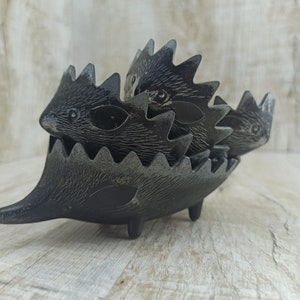 May include: A black metal hedgehog-shaped ashtray with four individual compartments. The ashtray is designed to look like a group of hedgehogs huddled together.