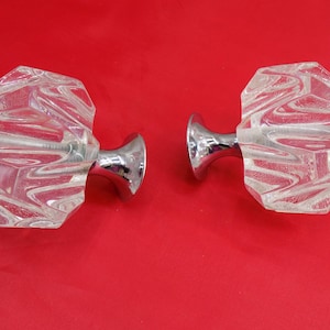 May include: Two clear glass cabinet knobs with faceted, octagonal shapes. The knobs have a silver-coloured metal base and a screw for mounting. The background is a solid red.